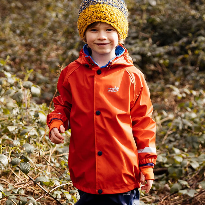 A boy wearing a red Rainy Day Waterproof Jacket standing in a wooded area smiling at the camera. He also has on a yellow and blue bobble hat.